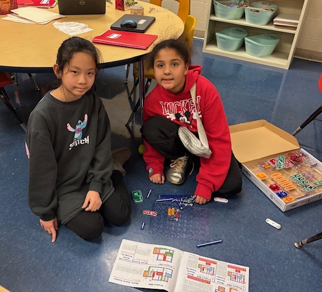 Two students sit on the floor working together on a Snap Circuits project, following instruction sheets while connecting electronic components on a circuit board.