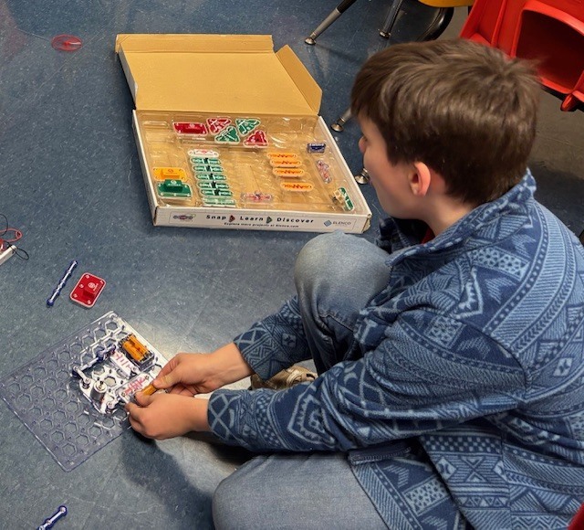 A student sits on the classroom floor assembling a Snap Circuits project, connecting electronic pieces on a clear grid board while an open Snap Circuits kit box sits nearby.