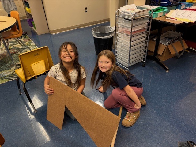 Two students kneel on the classroom floor next to a large cardboard aqueduct design piece, smiling and posing with their completed engineering project.