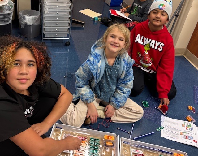 our students sit on the classroom floor surrounded by Snap Circuits kits and instruction pages, collaborating to build electronic circuits during a STEM activity.