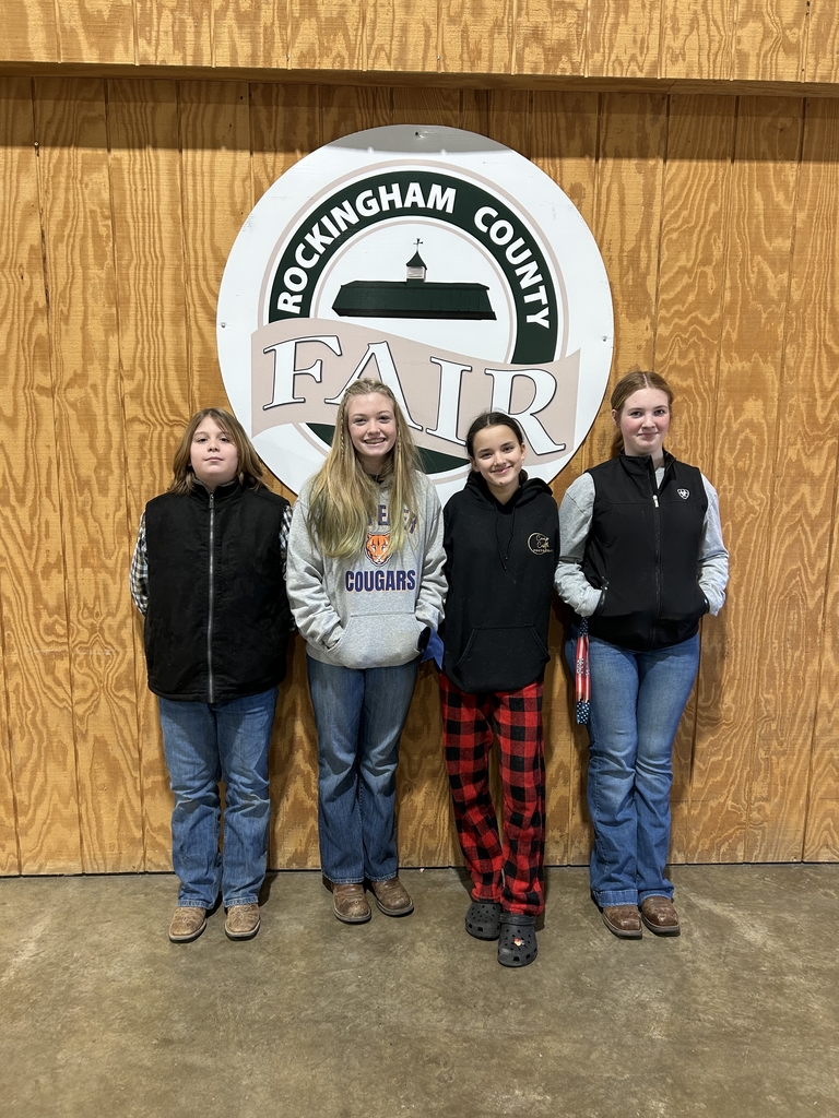 Four middle school students stand side by side indoors in front of a wooden wall with a large circular sign that reads “Rockingham County Fair.” The students are wearing casual clothing including jeans, boots, and jackets or hoodies, and they are smiling at the camera. The floor is concrete, and the setting appears to be inside a livestock or fair facility.