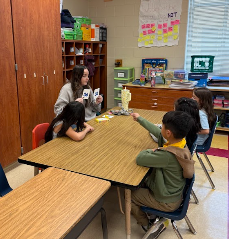 A teacher sits at a table holding up a number card while four young students sit around the table participating in a small-group math activity.