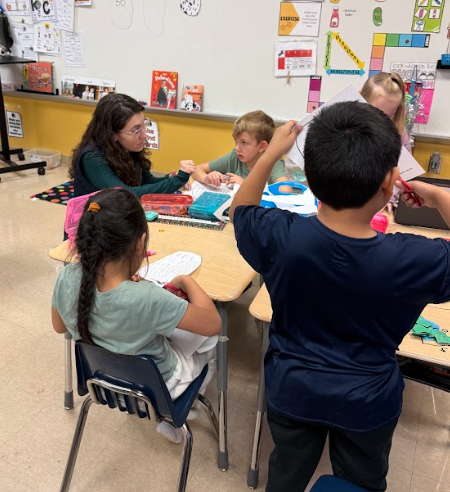 A small group of elementary students sit around a table working on a classroom activity while an adult helper assists and observes nearby.