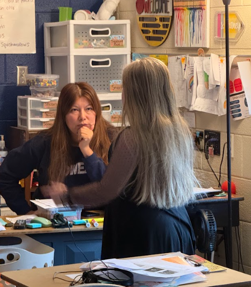 One  adult and one student stand facing each other at a teacher’s desk engaged in conversation, with classroom supplies, papers, and posters visible in the background.