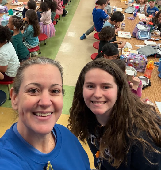 Two smiling adults take a selfie in a busy school cafeteria while elementary students sit at long tables eating lunch behind them.