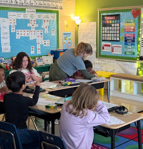 A teacher assists a group of elementary students at desks while they complete assignments in a brightly decorated classroom.