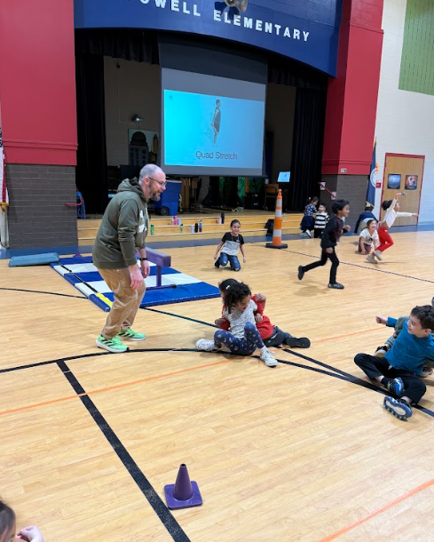 A PE teacher supervises students participating in an indoor gym activity, with children running, stretching, and sitting on mats in a school gymnasium.