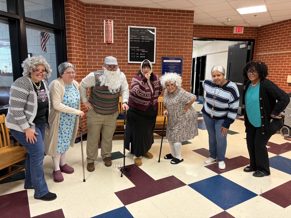 Eight adults stand in a school hallway wearing gray wigs and costumes to look like elderly people, some using canes and walkers. They pose playfully in front of a brick wall and bench, smiling and leaning forward, as if participating in a themed spirit day or skit.