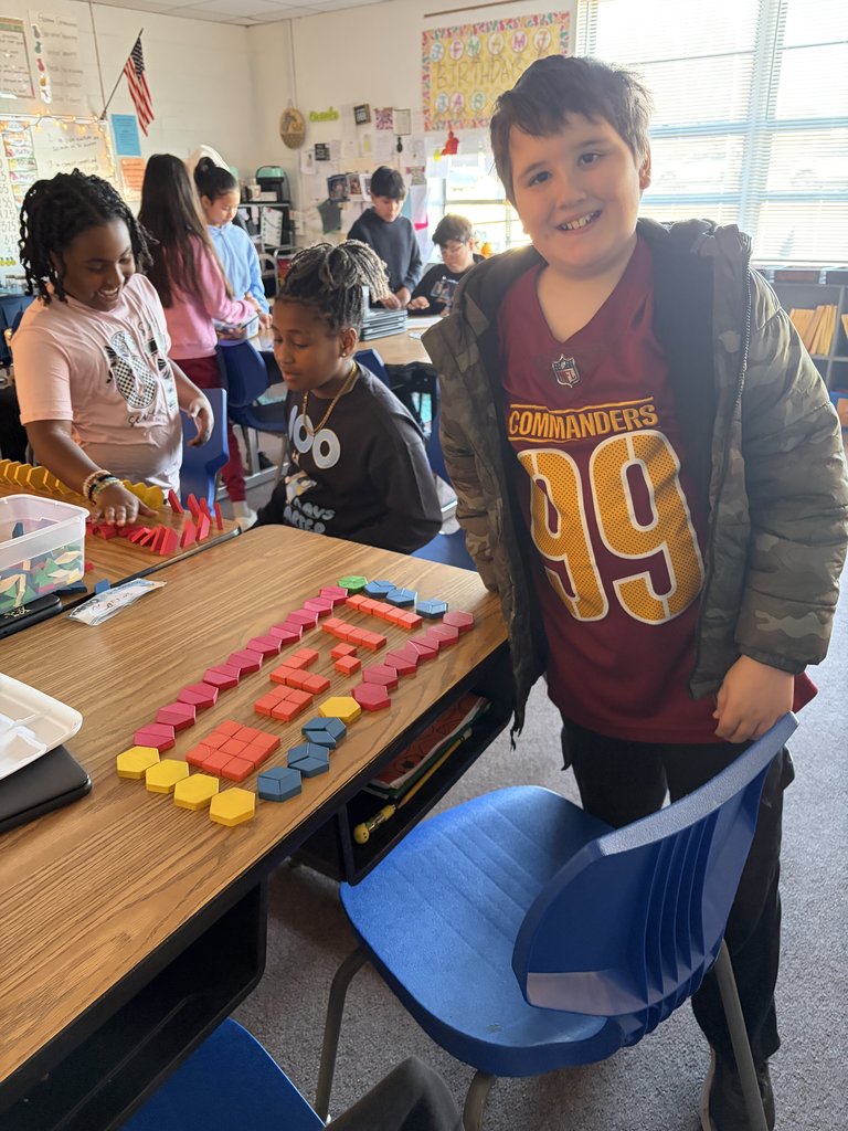 A young boy in a maroon Washington Commanders jersey smiles next to a desk where a pattern of red, yellow, and blue geometric blocks has been carefully arranged.