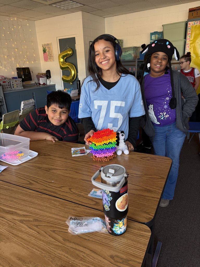 Three smiling students stand behind a desk showcasing a tall, tiered tower made of colorful rainbow-patterned beads. A small stuffed panda sits next to the tower, and a personalized water bottle is in the foreground.
