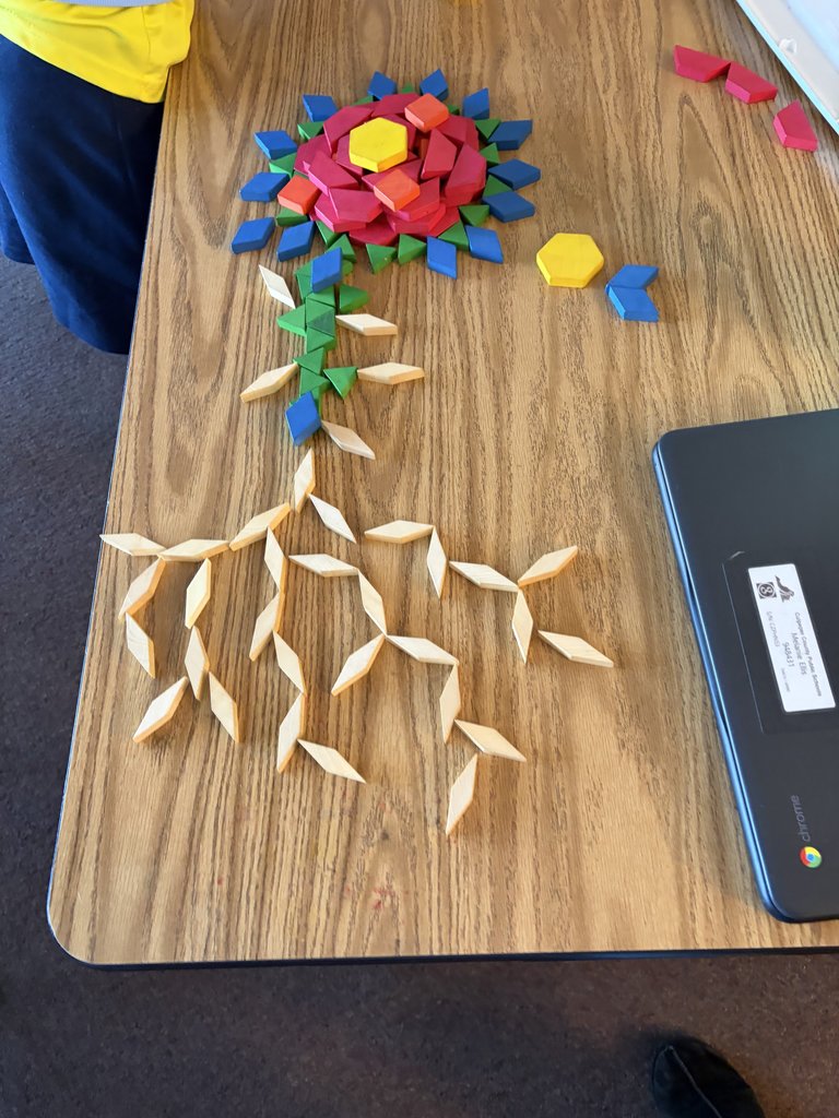 A high-angle shot of an intricate flower design made from pattern blocks on a desk, featuring a complex root system made of tan diamond-shaped blocks and a blue laptop sitting nearby.