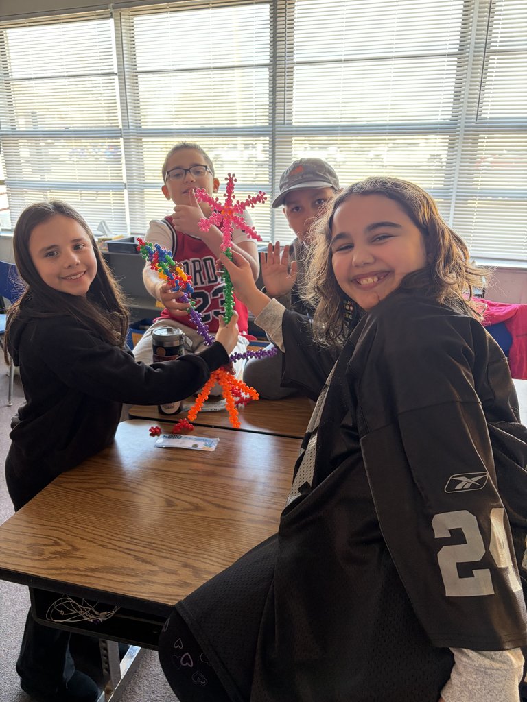 Four students pose together at a desk, proudly holding up a tall, branching structure they built using colorful interlocking plastic star-shaped pieces.