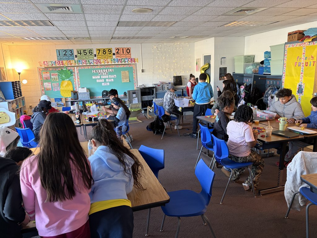 A bright, busy classroom filled with elementary students working in small groups at wooden desks. The walls are decorated with educational posters, including a large "Invisible Math" board and a colorful number line.