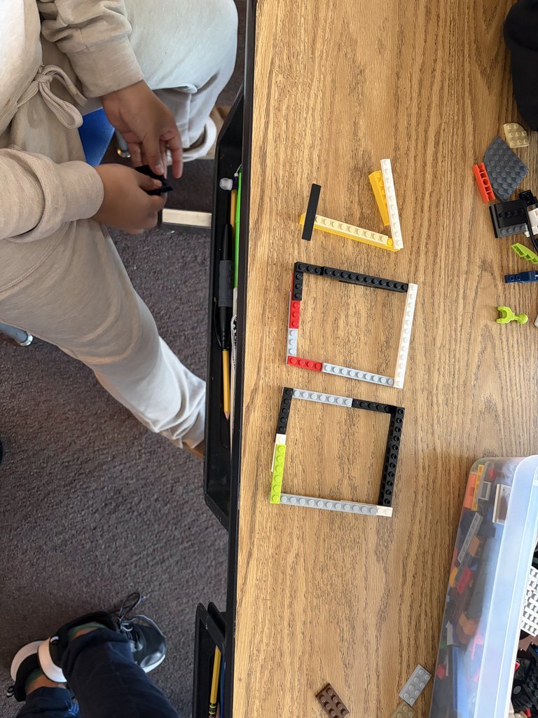 A top-down view of a student using colorful LEGO bricks on a wooden desk to construct the number "100."