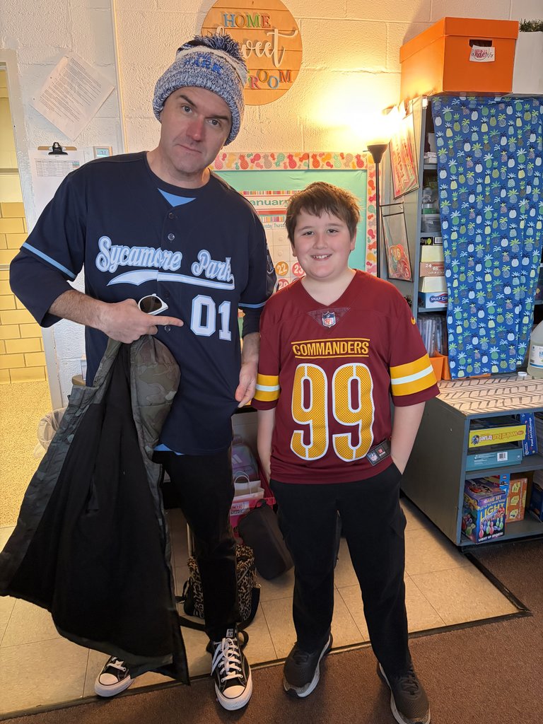 A man in a blue "Sycamore Park" jersey and a blue knit beanie stands next to a smiling young boy in a maroon "Commanders" jersey inside a classroom.