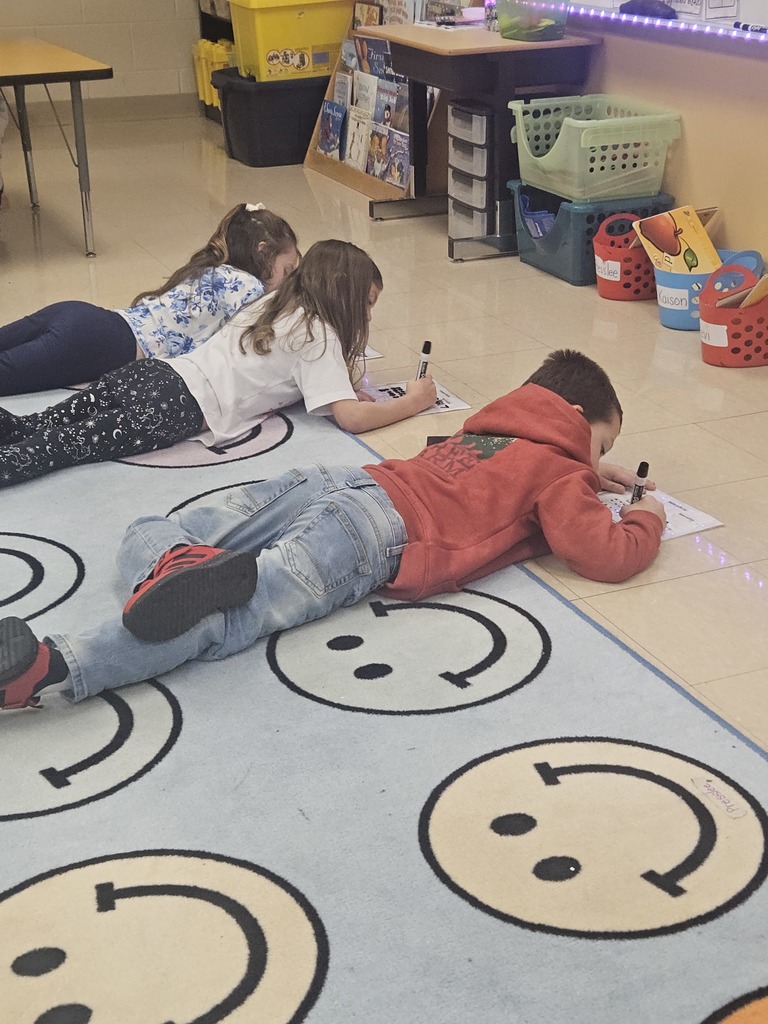 Three elementary students lie on a classroom rug while writing on dry-erase math worksheets with markers, working quietly during a floor activity.