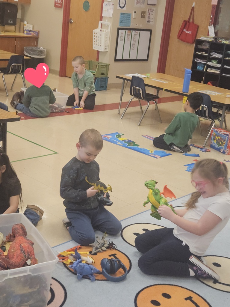 Several students sit on a rug playing with dinosaur toys and figures during center time, smiling and interacting with classmates.