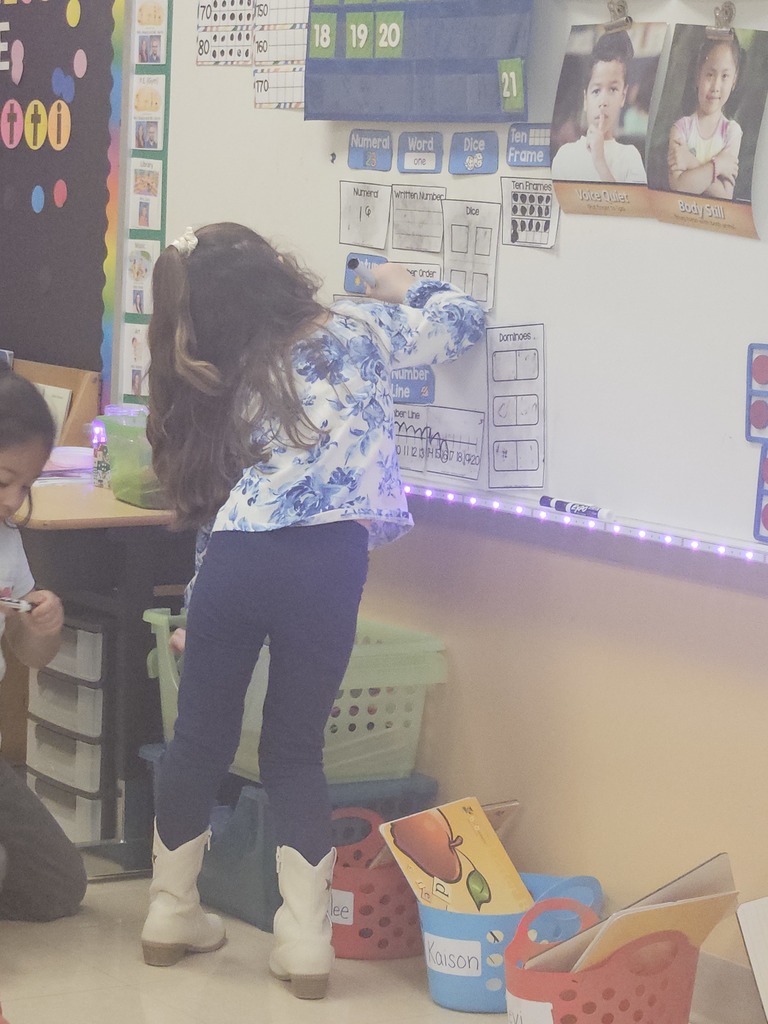 A student stands at a whiteboard using a dry-erase marker to complete a number activity chart during a math station.