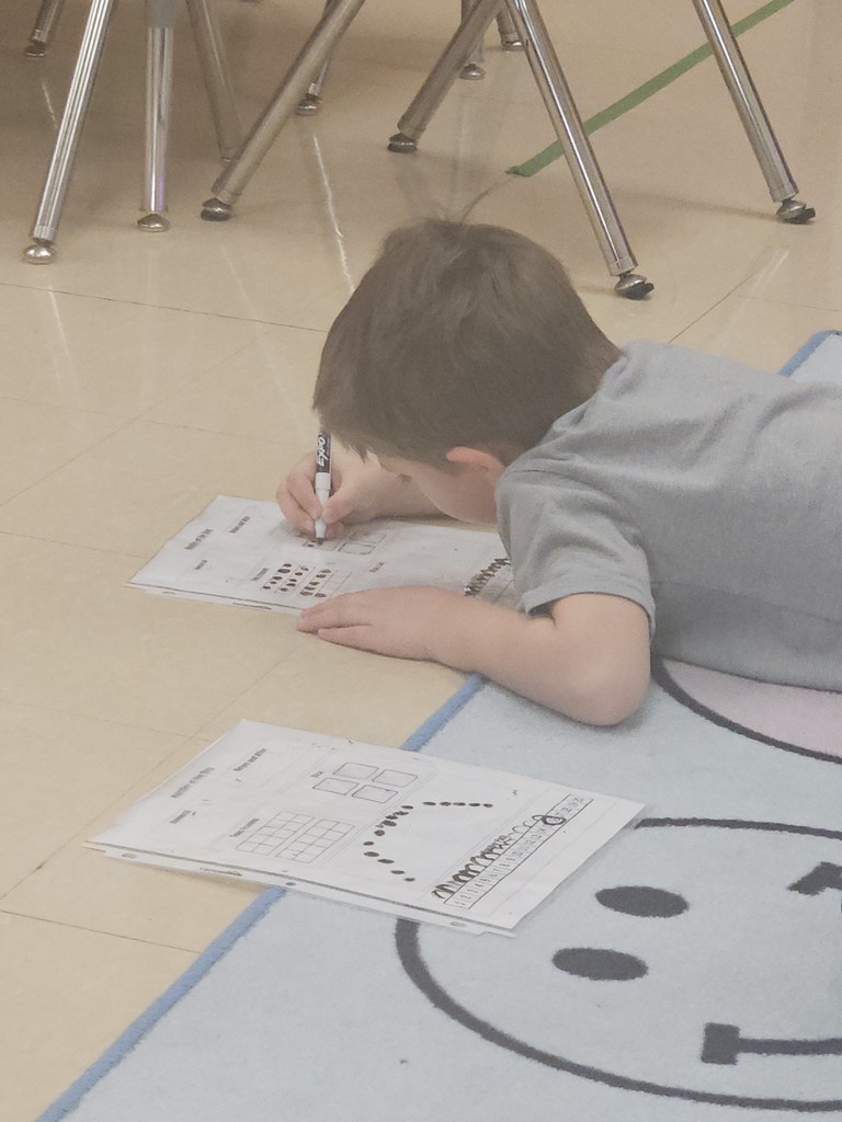 A student lies on the rug writing answers on a laminated math worksheet using a dry-erase marker.