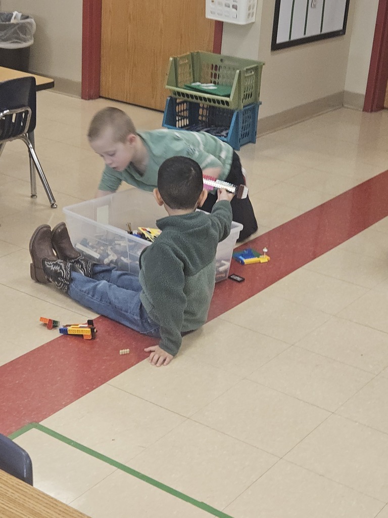 Two students sit on the classroom floor sorting and building with small manipulatives from a clear plastic bin.