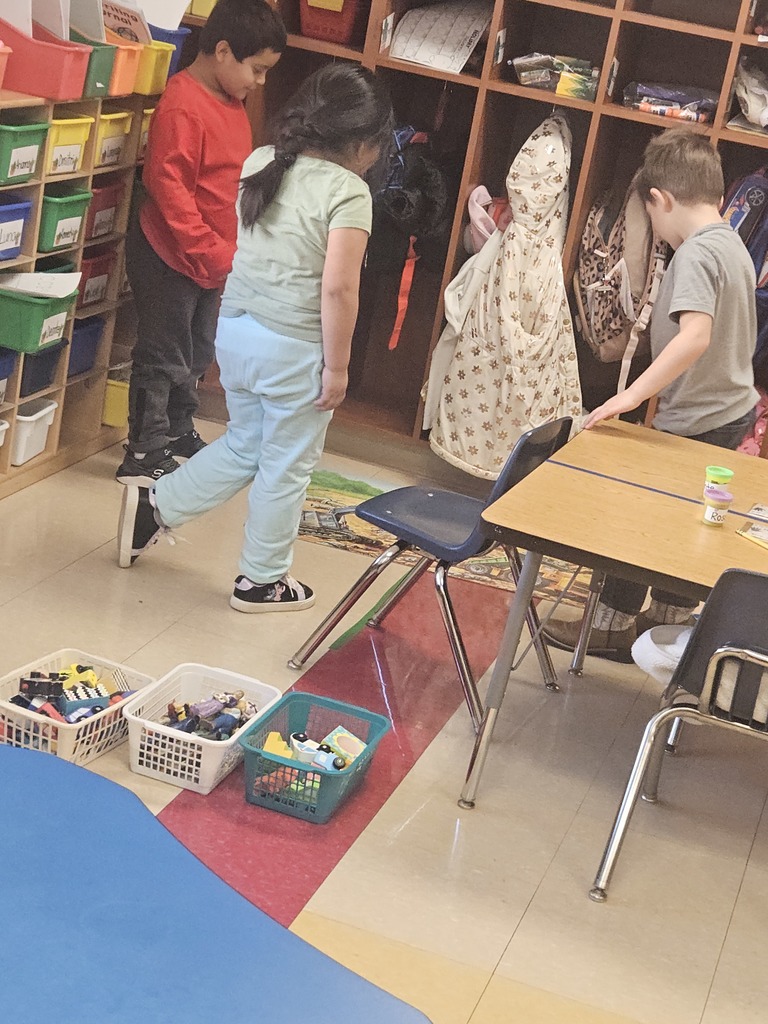 Students stand near cubbies and desks while organizing materials and transitioning between classroom activities.