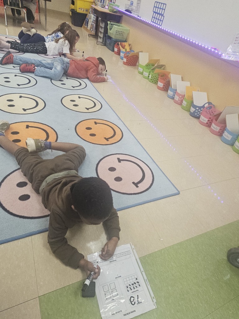 Three students lie on the rug working independently on number line math worksheets with dry-erase markers.