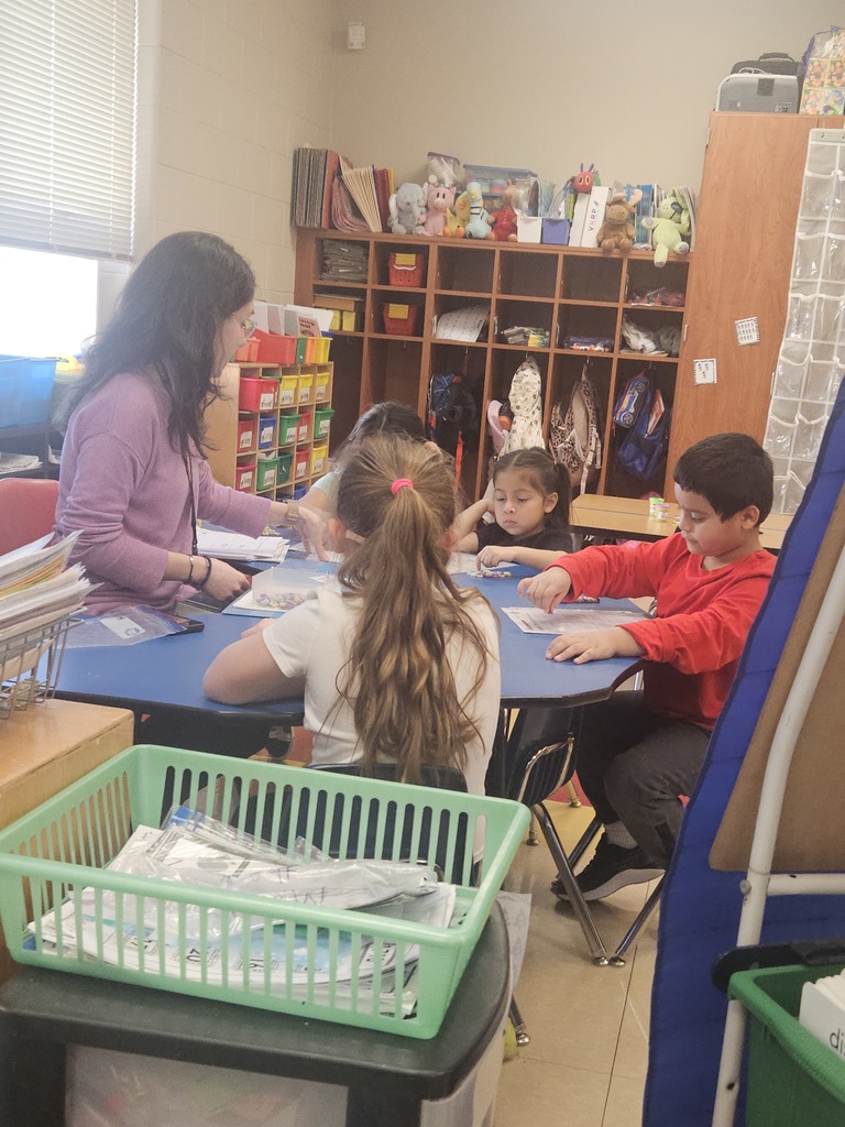 A teacher sits at a small table working with a small group of students during guided instruction, assisting with hands-on learning materials.