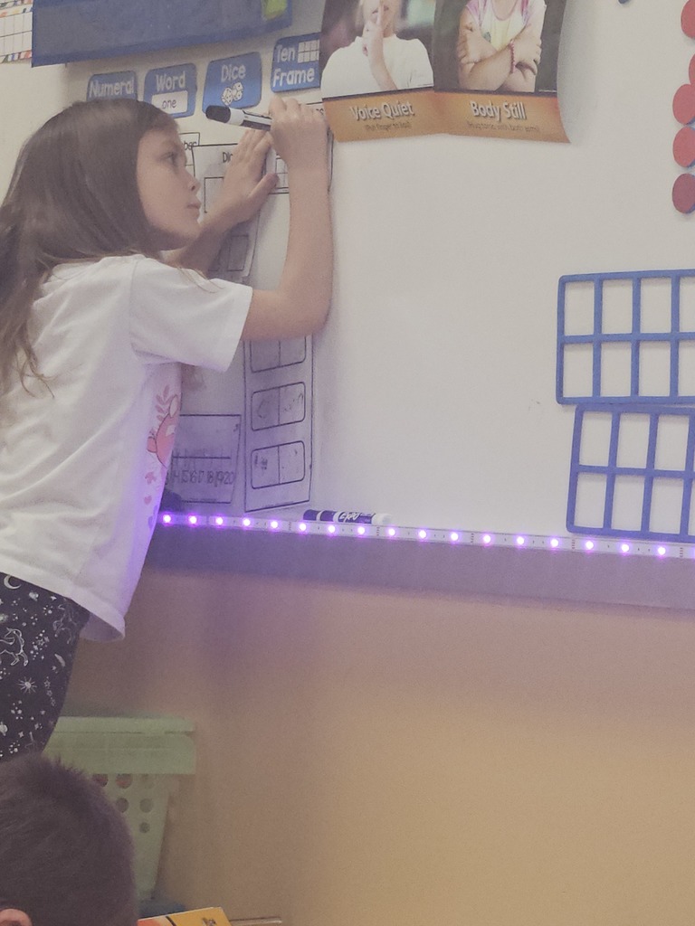 A student stands at the classroom whiteboard writing on a math chart under visual behavior reminders such as “Voice Quiet” and “Body Still.”