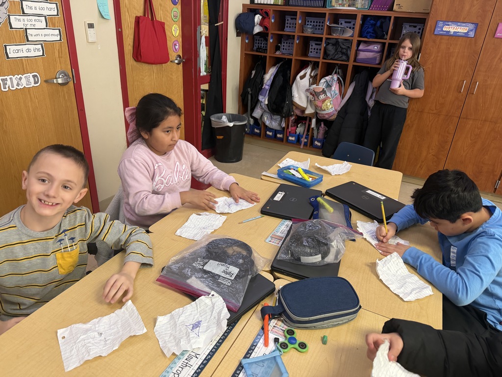 A small group of elementary students sit around a table writing and folding paper during a math activity, with Chromebooks, pencils, and supplies spread across the tabletop.