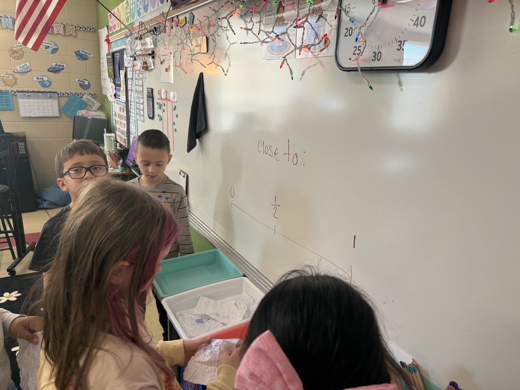 Several students stand near a classroom whiteboard placing paper responses into colored bins while reviewing answers during a whole-group math activity.