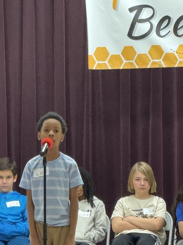 A student stands at a microphone on a stage during a spelling bee with other contestants seated in a row behind him.