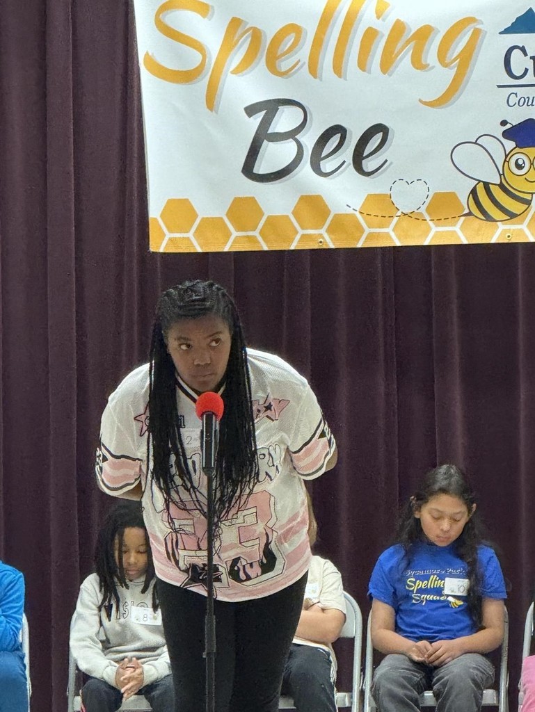 A student stands at a microphone on a stage during a spelling bee while other contestants sit behind her beneath a “Spelling Bee” banner.