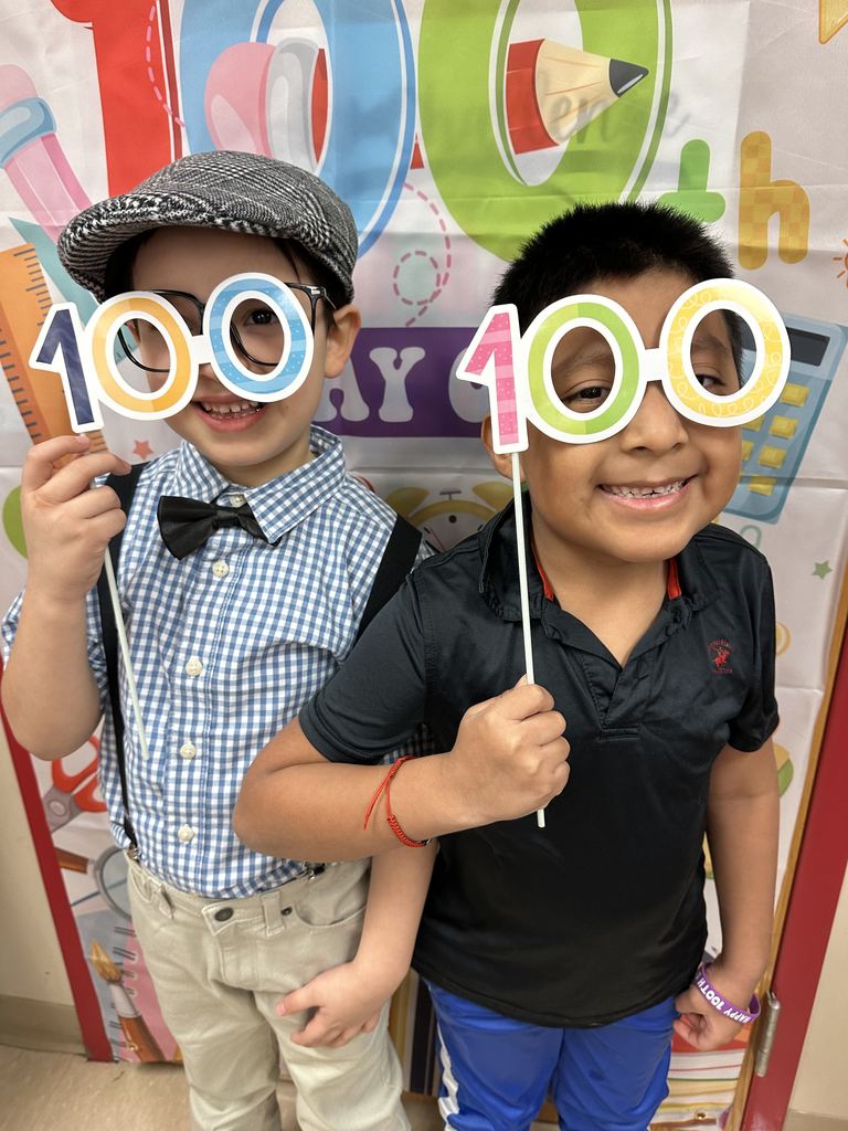 Two smiling students pose in front of a colorful “100th Day” backdrop while holding oversized “100” glasses on sticks.