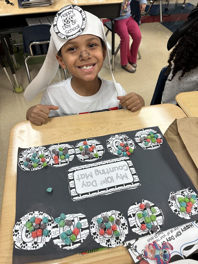 A smiling student sits at a desk wearing a “Happy 101st Day of School” hat and displays a counting mat filled with colorful cereal pieces.