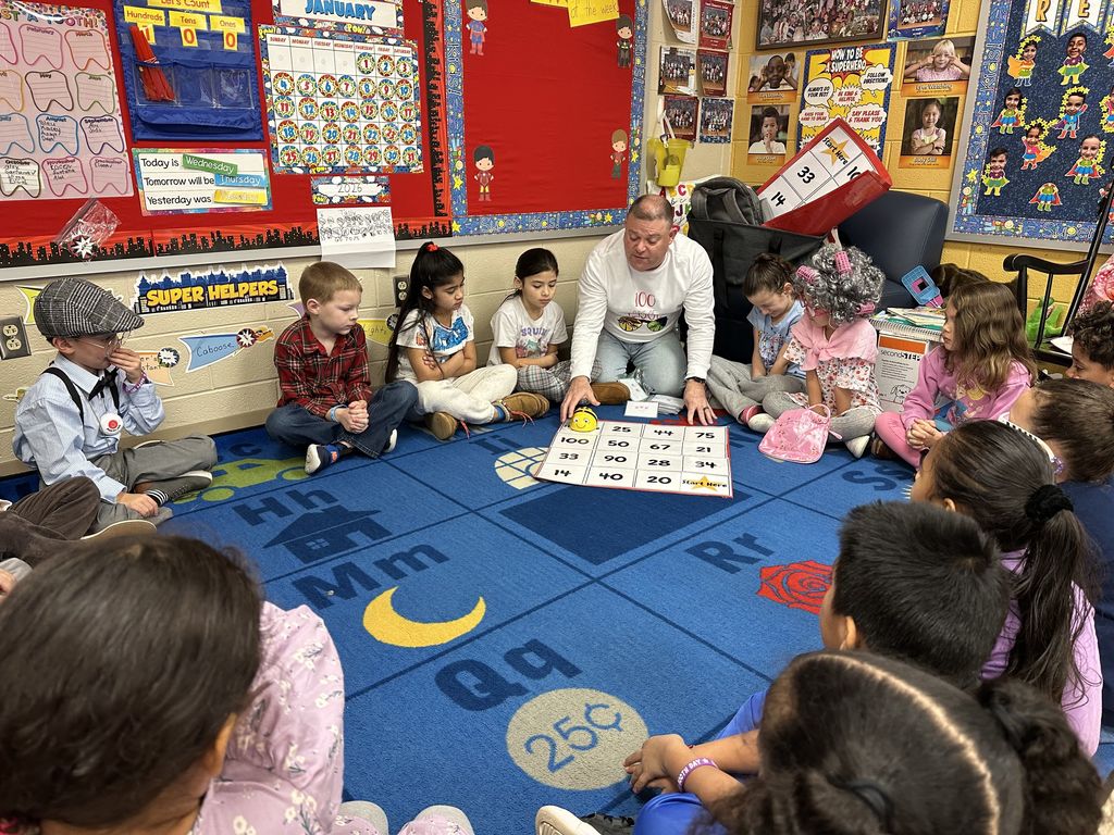 Students sit in a circle on a blue classroom rug while a teacher leads a 100th Day of School counting game using a large number mat in the center.