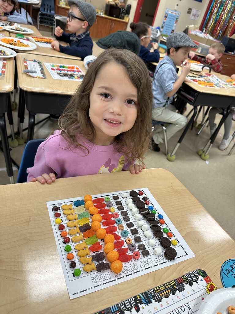 A student sits at a desk smiling at the camera with a completed 100 chart filled with assorted snacks arranged in rows.