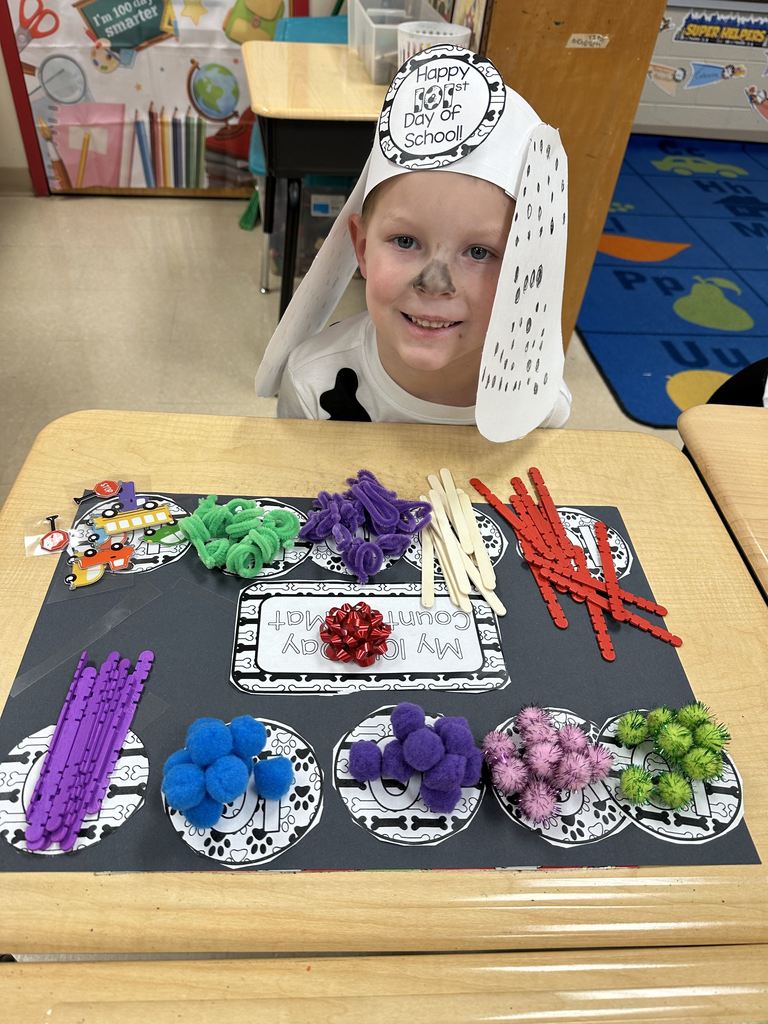 A student wearing a 101st Day of School hat smiles at the camera while showing a counting mat with grouped classroom materials such as pom-poms and craft sticks.