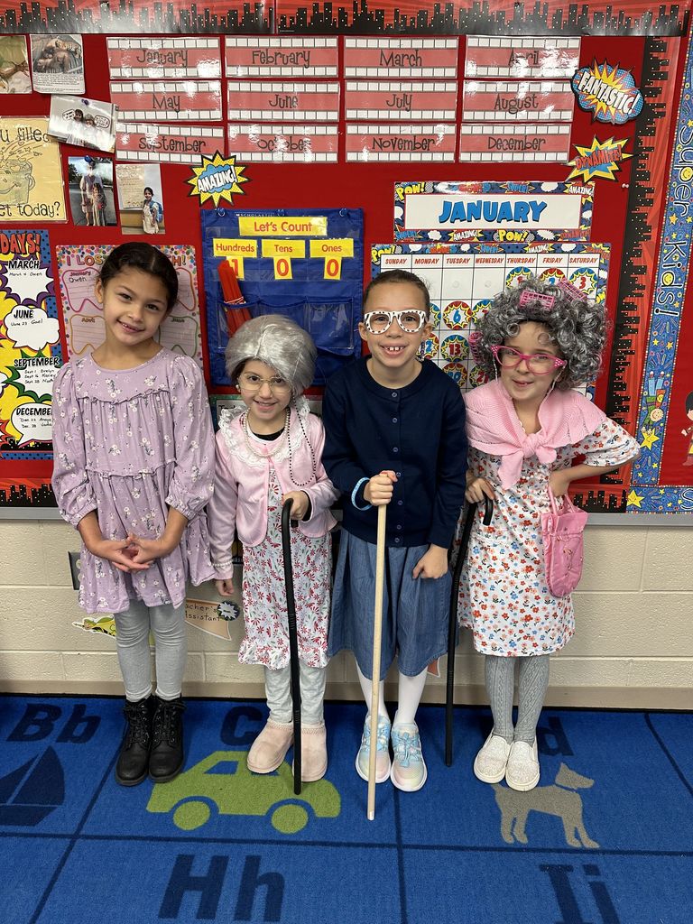 Four students dressed as “100-year-old” characters pose in front of a classroom calendar board, wearing glasses, shawls, and holding canes.