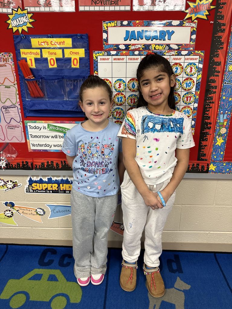 Two students stand together smiling in front of a classroom calendar display while wearing shirts decorated for the 100th Day of School.