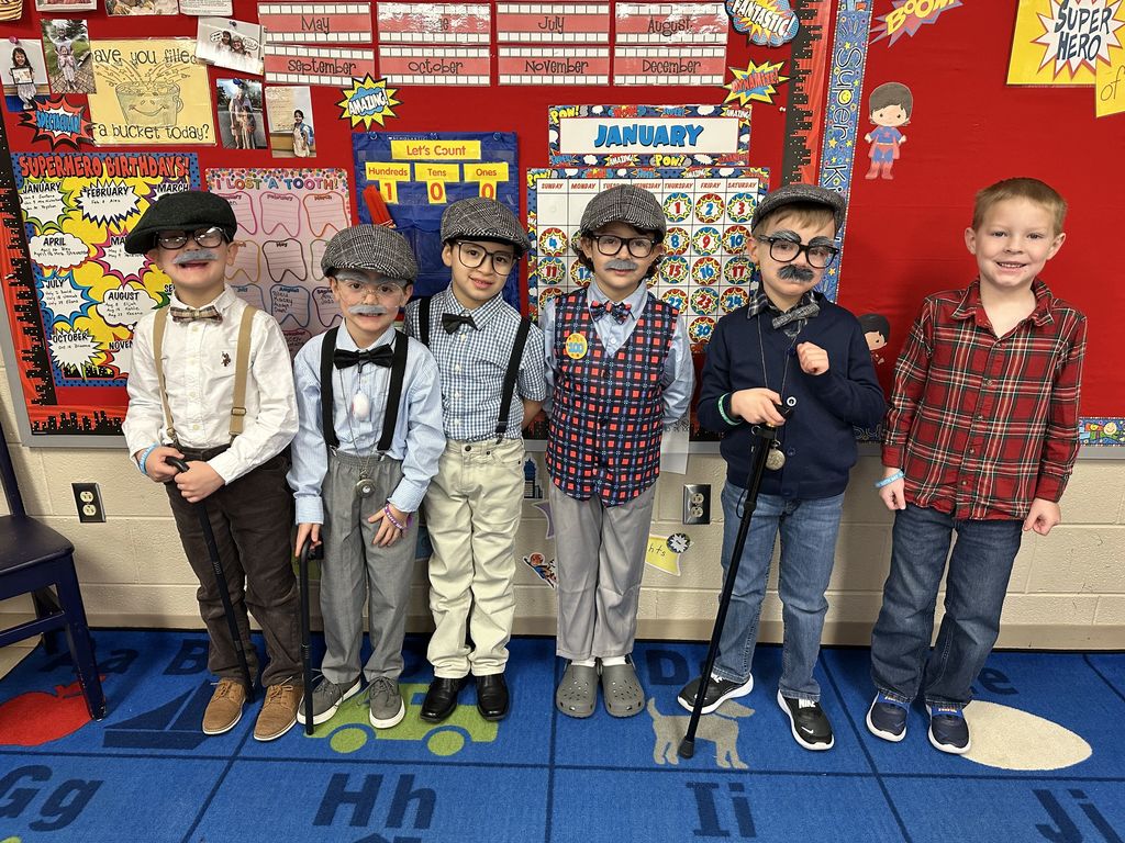 A group of students dressed as “100-year-old” characters pose in a line wearing hats, suspenders, glasses, fake mustaches, and holding canes.