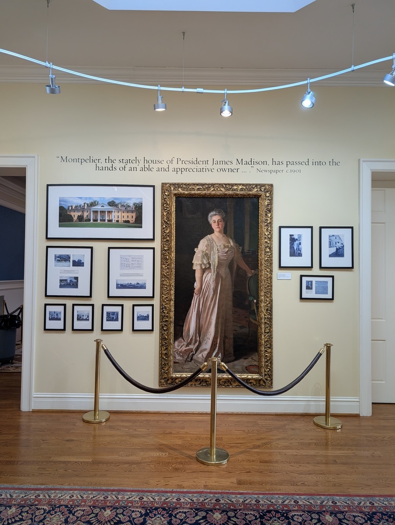 An indoor museum exhibit wall featuring a large, gold-framed oil portrait of a woman in a formal, floor-length pink gown. Surrounding the portrait are several smaller framed photographs and documents. A quote printed on the wall above reads, “Montpelier, the stately house of President James Madison, has passed into the hands of an able and appreciative owner...” Gold stanchions with a dark rope barrier stand in front of the display.