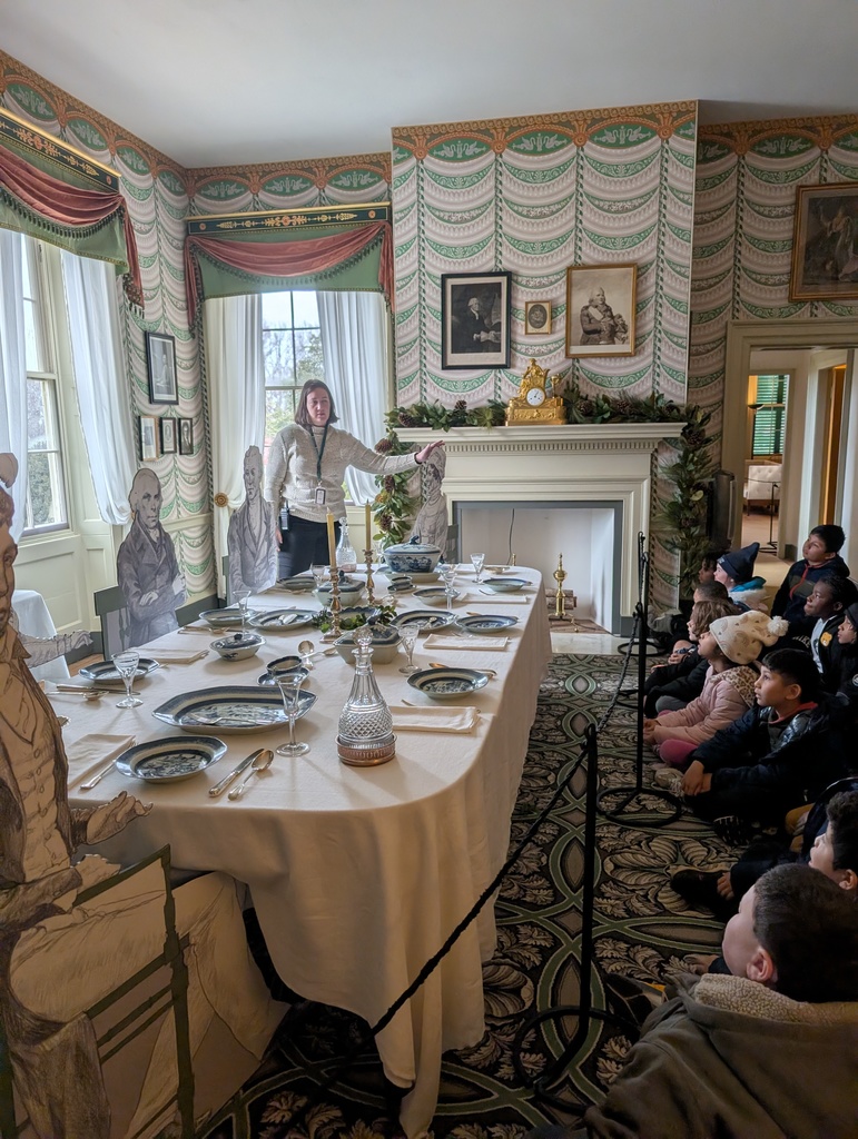 A wide view of a large, formal room with high ceilings, an ornate gold chandelier, and a decorative plaster ceiling medallion. The room features dark blue walls with white wainscoting and three large windows draped in heavy gold curtains. A massive, circular patterned rug in brown, cream, and light blue covers most of the polished wood floor. In the background, a fireplace is decorated for the holidays with a small Christmas tree and greenery.
