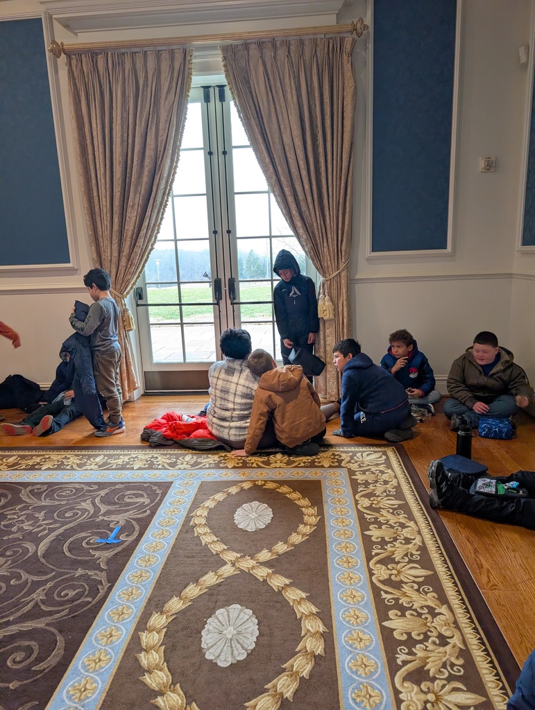A group of children in winter coats and casual clothes are sitting and standing on a wooden floor in a grand room. They are gathered in front of large glass French doors that look out onto a grassy landscape. To the sides of the doors are heavy gold drapes, and a portion of a large, ornate brown and cream rug is visible in the foreground.