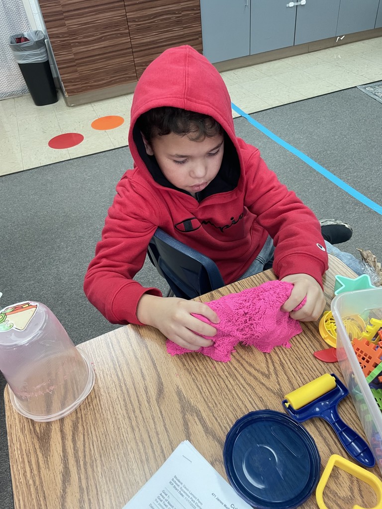 A boy wearing a red hoodie sits at a desk, focusing intently on squeezing and molding a large pile of bright pink kinetic sand. Various sand tools and a yellow roller are scattered on the table around him.