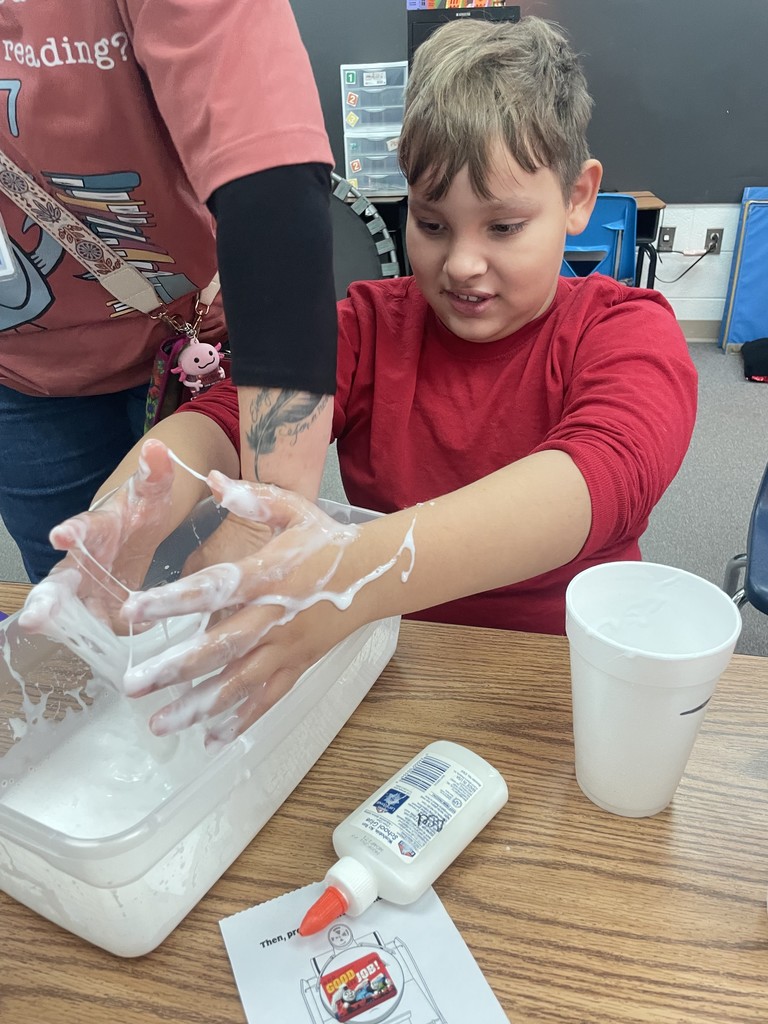 A young boy in a red shirt reaches into a clear bin filled with stretchy white slime. His hands are covered in the gooey substance, which strings between his fingers as an adult assists him. A bottle of glue and a "Good Job" sticker are on the table.