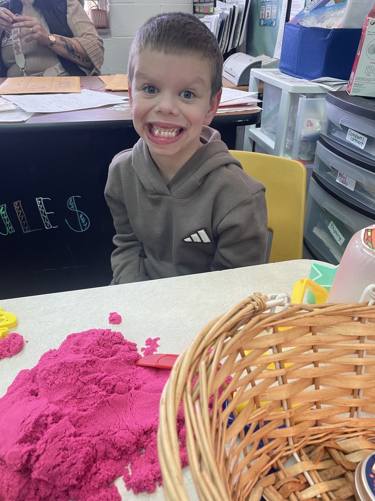 A young boy in a brown Adidas hoodie looks at the camera with a wide, excited grin. On the table in front of him is a large mound of bright pink kinetic sand and a wicker basket.
