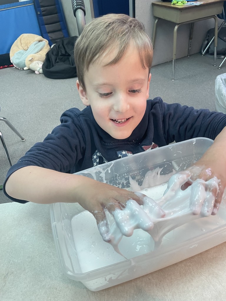 A young boy in a dark blue hoodie smiles joyfully as he pulls and stretches a thick, white substance in a clear plastic container during a sensory play activity in a classroom.
