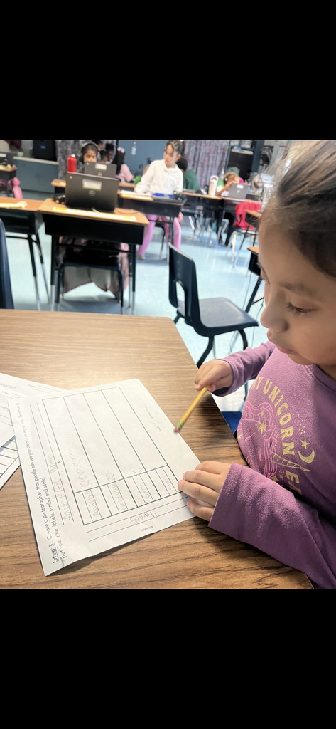 A student in a purple "Unicorn" sweatshirt works on a "Favorite Games" pictograph. She is using a yellow pencil to draw heart symbols in the categories for "Roblox," "Bouncy," and "Tag." Other students with laptops are visible in the background.
