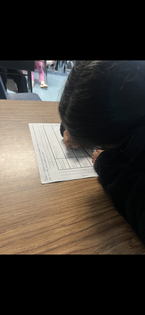 A close-up, high-angle shot of a student with dark hair leaning over a desk, diligently working on a pictograph. The worksheet is titled "Favorite Food" and includes categories like pizza and hot dogs. The student is focused on drawing small symbols into the graph's columns.