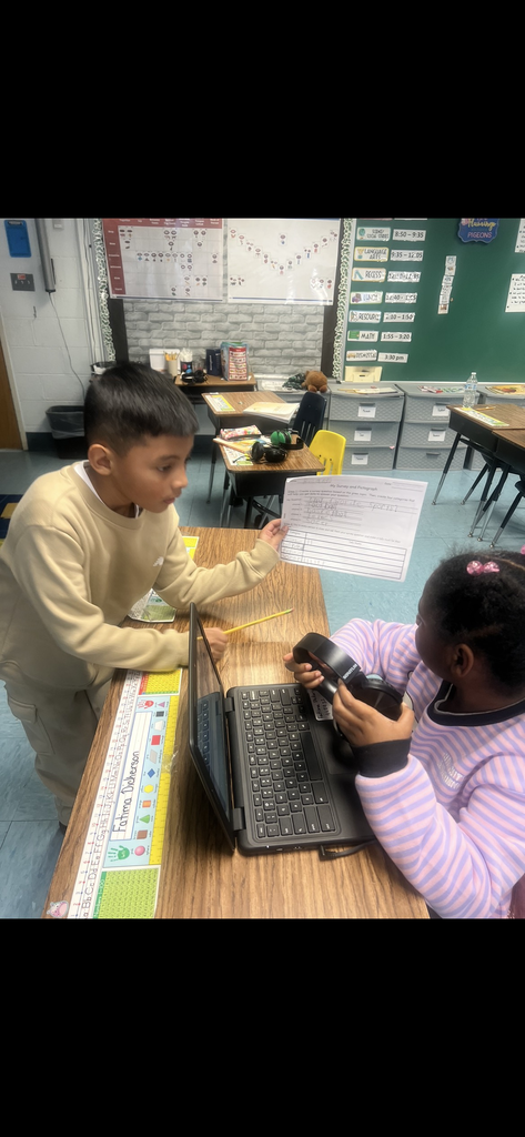 A young boy in a tan sweatshirt leans over a desk to show his "Favorite Sports" survey to a classmate. The classmate, wearing a striped purple sweater and headphones, looks up from her laptop to see the paper. The worksheet lists football, basketball, tennis, and soccer.